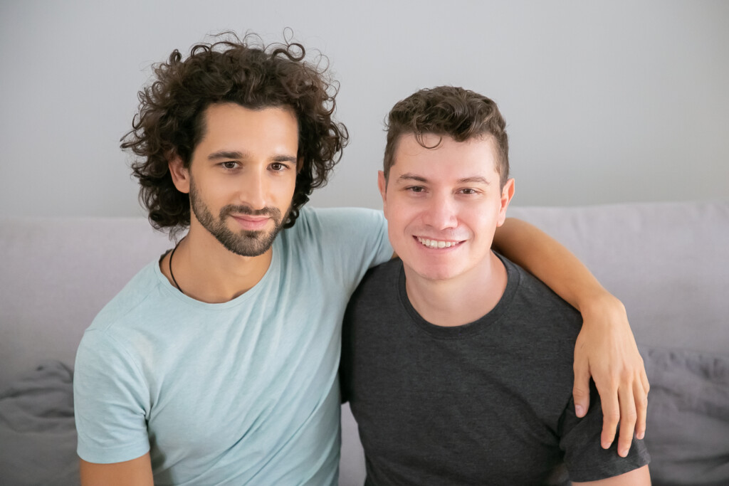 Happy handsome gay couple posing at home, sitting on couch together and hugging each other. Front view. Love and relationships concept